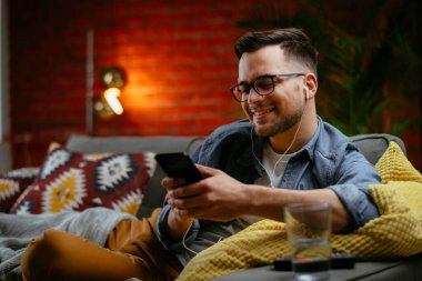 Genç adam kanepede uzanıp müzik dinliyor. Modern adam oturma odasında telefonuyla müzik dinliyor..