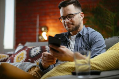 Genç adam kanepede uzanıp müzik dinliyor. Modern adam oturma odasında telefonuyla müzik dinliyor..