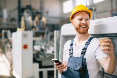 Sarı miğferli mutlu işçi fabrikada telefon kullanıyor. 