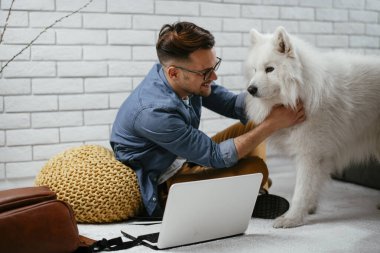 Adam dizüstü bilgisayarla çalışırken köpeğiyle yerde oynuyor. Samoe köpeğiyle oynayan adam..