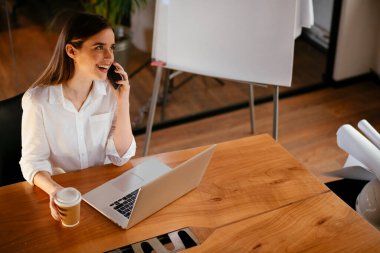 İş kadını ofiste dizüstü bilgisayar kullanıyor ve telefonla konuşuyor.