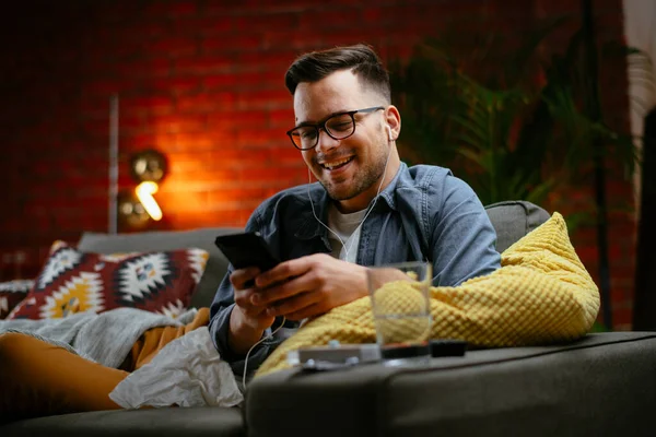 Genç adam kanepede uzanıp müzik dinliyor. Modern adam oturma odasında telefonuyla müzik dinliyor..