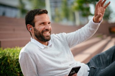 Dışarıdaki yakışıklı iş adamının portresi. Kulaklıklı genç adam dışarıda oturuyor ve telefon kullanıyor..