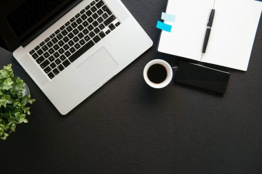 Ofis masasının en üst görüntüsü. Laptop, kahve ve malzeme dolu siyah ofis masası..