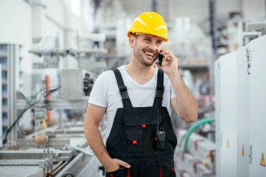 Sarı kasklı yakışıklı işçi fabrikada telefon kullanıyor.