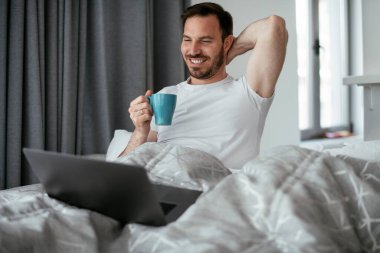 Genç adam yatakta film izliyor ve kahve içiyor. Yakışıklı adam sabahları eğleniyor..