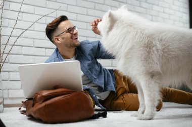 Adam dizüstü bilgisayarla çalışırken köpeğiyle yerde oynuyor. Samoe köpeğiyle oynayan adam..