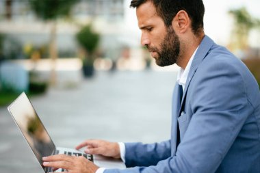 Mavi takım elbiseli iş adamı dışarıda dizüstü bilgisayarında yazı yazıyor.