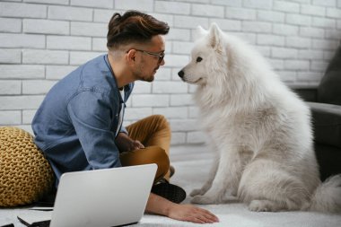 Adam dizüstü bilgisayarla çalışırken köpeğiyle yerde oynuyor. Samoe köpeğiyle oynayan adam..