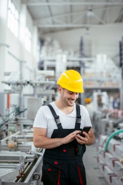 Sarı kasklı yakışıklı işçi fabrikada telefon kullanıyor.