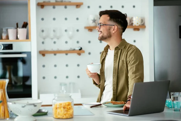 Genç iş adamı evde dizüstü bilgisayarıyla çalışıyor. Evde kahvaltı yaparken çalışan yakışıklı adam..