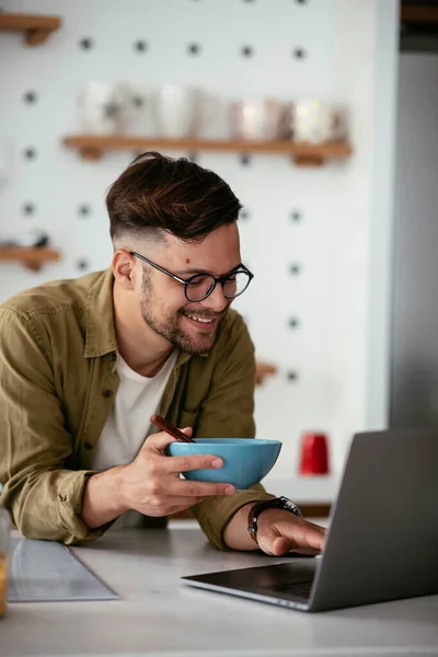 Young businessman working from home on his laptop. Handsome man working while eating breakfast at home.