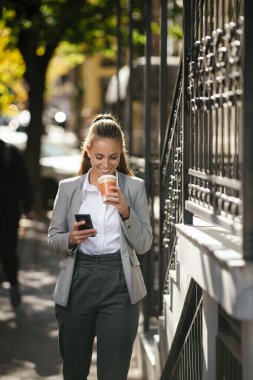 Genç ve güzel iş kadını sokakta yürürken kahve molasında telefon kullanıyor.