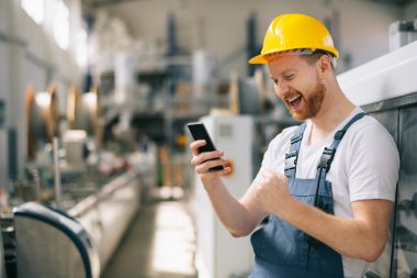 Sarı miğferli mutlu işçi fabrikada telefon kullanıyor. 
