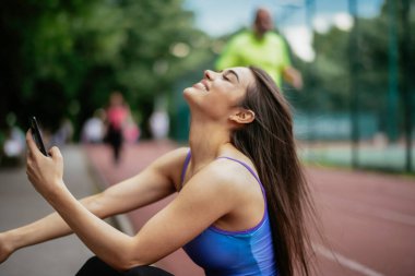 Egzersiz yapmaktan dinlenen güzel bir kadın. Genç sporcu kadın bankta oturuyor ve telefonu kullanıyor..