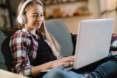 Şirin kız kulaklıkla müzik dinliyor. Dizüstü bilgisayarla kanepede oturan küçük kız..