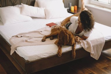 Güzel genç bir kadın köpeğiyle yatakta oynuyor.