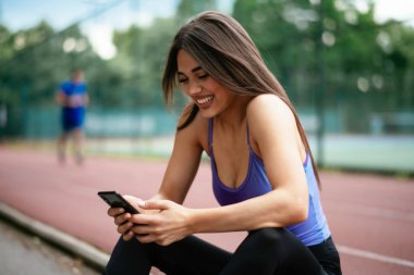 Egzersiz yapmaktan dinlenen güzel bir kadın. Genç sporcu kadın bankta oturuyor ve telefonu kullanıyor..