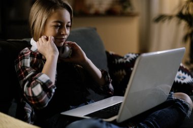 Şirin kız kulaklıkla müzik dinliyor. Dizüstü bilgisayarla kanepede oturan küçük kız..