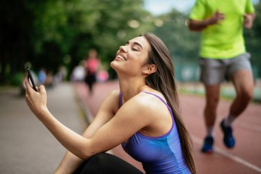 Egzersiz yapmaktan dinlenen güzel bir kadın. Genç sporcu kadın bankta oturuyor ve telefonu kullanıyor..