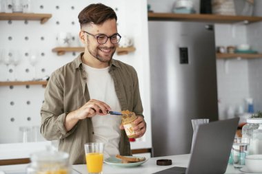 Genç adam kahvaltı yapıyor ve internette haberleri okuyor. Yakışıklı adam evde eğleniyor..