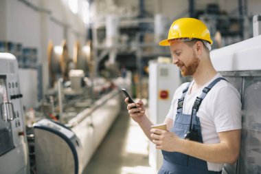 Sarı miğferli mutlu işçi fabrikada telefon kullanıyor. 