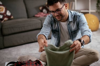 Giysilerini seyahat çantasına koyan gülümseyen genç adam. Yolculuğa hazırlanan adam.
