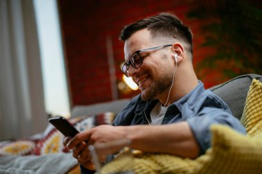 Genç adam kanepede uzanıp müzik dinliyor. Modern adam oturma odasında telefonuyla müzik dinliyor..