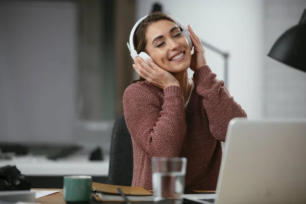 Young woman in office with headphones. Young casual businesswoman enjoying in her favorite song over headphones and singing while working on laptop in the office.