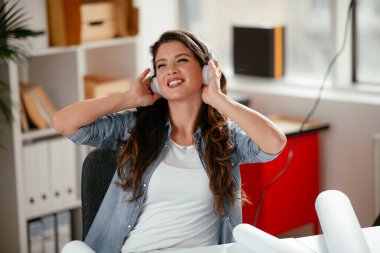 Ofiste kulaklıklı genç bir kadın. Genç gündelik iş kadını en sevdiği şarkıyı kulaklıkla dinliyor ve ofiste dizüstü bilgisayarla çalışırken şarkı söylüyor..