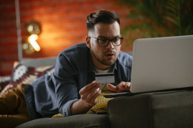 Genç adam oturma odasında online alışveriş için kredi kartı kullanıyor..