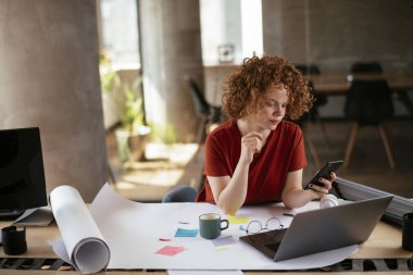 Güzel bir iş kadını proje üzerinde çalışıyor. Ofiste akıllı telefon kullanan kıvırcık saçlı genç bir kadın..