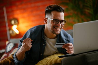 Genç adam oturma odasında online alışveriş için kredi kartı kullanıyor..