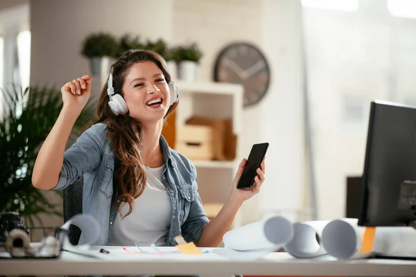 Young woman in office with headphones. Young casual businesswoman enjoying in her favorite song over headphones and singing while working on laptop in the office.