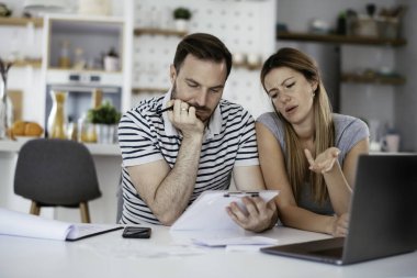 Yorgun çift mutfaktaki masada belgeleri inceliyor.