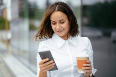 Sokakta telefon ve kahveyle yürüyen genç bir kadın.