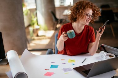 Güzel bir iş kadını proje üzerinde çalışıyor. Ofiste akıllı telefon kullanan kıvırcık saçlı genç bir kadın..