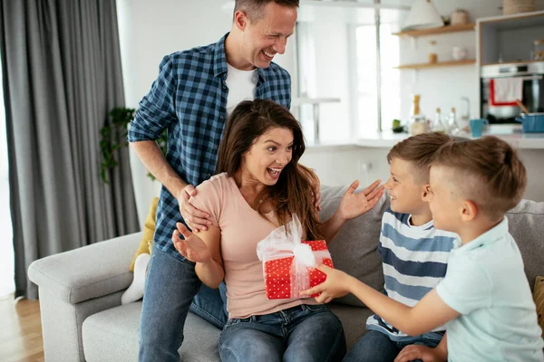 Father with two young sons are giving their mother a gift. Mother is surprised to receive a present from sons and husband