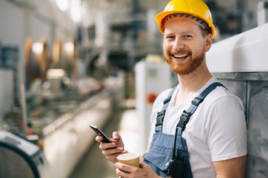 Sarı miğferli mutlu işçi fabrikada telefon kullanıyor. 