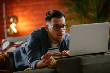 Genç adam oturma odasında online alışveriş için kredi kartı kullanıyor..