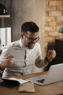 Gözlüklü ve beyaz gömlekli genç adam çalışma masasında dizüstü bilgisayarlı.