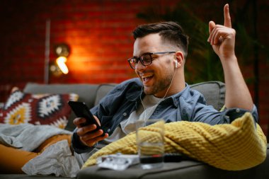 Genç adam kanepede uzanıp müzik dinliyor. Modern adam oturma odasında telefonuyla müzik dinliyor..