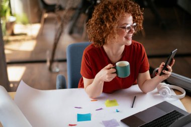 Güzel bir iş kadını proje üzerinde çalışıyor. Ofiste akıllı telefon kullanan kıvırcık saçlı genç bir kadın..