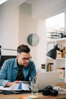 Çalışma masasında gözlüklü genç bir adam elinde kağıt rengi örnekleriyle.