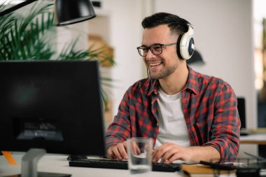 İş masasında kulaklık takan gözlüklü ve kareli gömlekli genç bir adam.