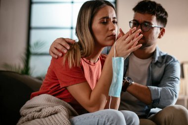 Tıbbi maskeli genç bir kadın ve erkek arkadaşı oturma odasında oturuyor.