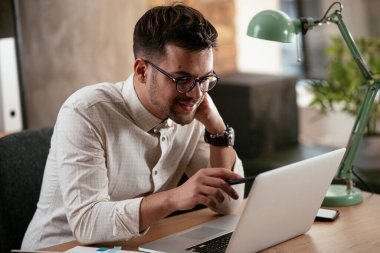 Gözlüklü ve beyaz gömlekli genç adam çalışma masasında dizüstü bilgisayarlı.