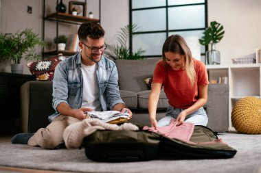 Genç adam ve kız arkadaşı oturma odasında bavul hazırlıyorlar.