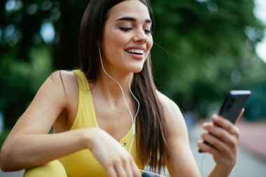 Egzersiz yapmaktan dinlenen güzel bir kadın. Genç sporcu kadın bankta oturuyor ve telefonu kullanıyor..