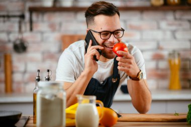 Önlük giyen genç adam mutfakta telefonla konuşuyor.